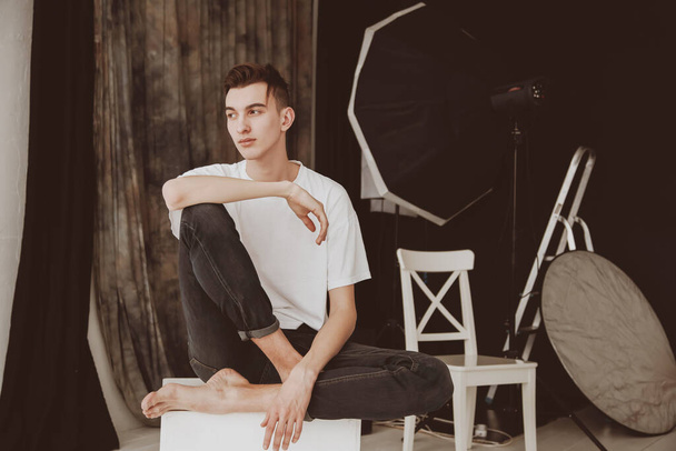A handsome male model, in a photo studio. On the background of studio equipment. A guy barefoot, in jeans and a white shirt. Sits on the cube. - Foto, Bild