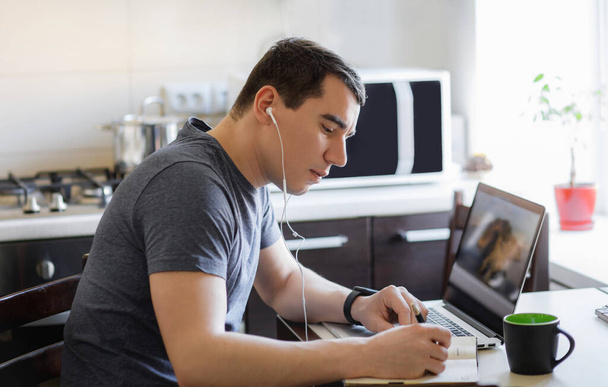 Un jeune homme travaille à distance à la maison à un ordinateur dans la cuisine, feuilletant un journal intime
 - Photo, image