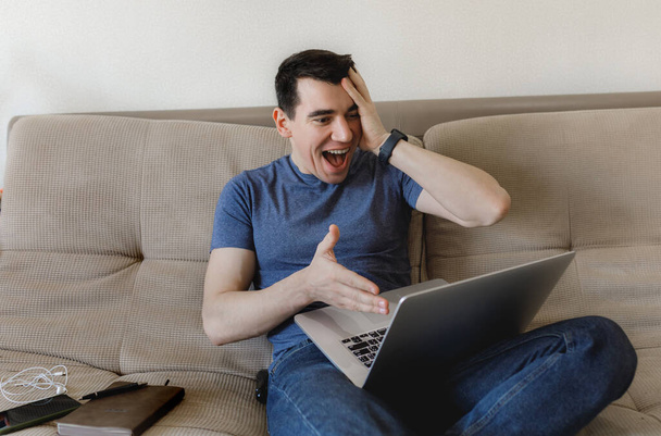 Молодой парень в синей футболке - европеец, сидящий дома на диване, работающий удаленно и общающийся с друзьями в интернете на ноутбуке с телефоном и дневником
 - Фото, изображение