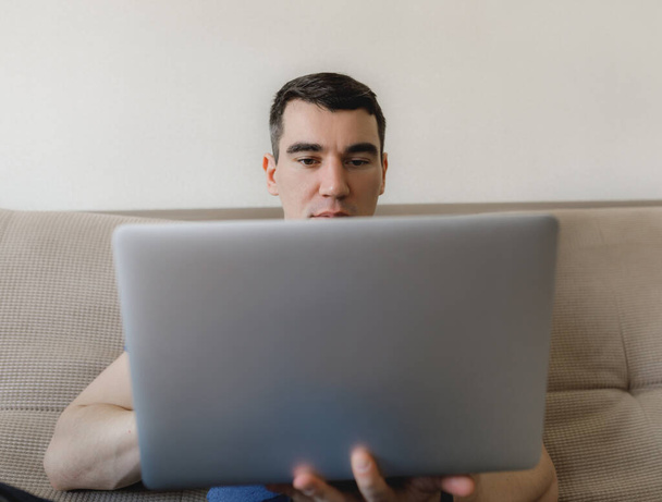 a young guy in a blue T-shirt is European sitting at home on the couch, working remotely and communicating with friends online on a notebook with a phone and a diary - Photo, Image