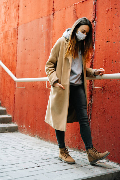uma jovem com um casaco castanho com uma máscara médica no rosto na rua, perto da parede vermelha. protecção contra o vírus
 - Foto, Imagem