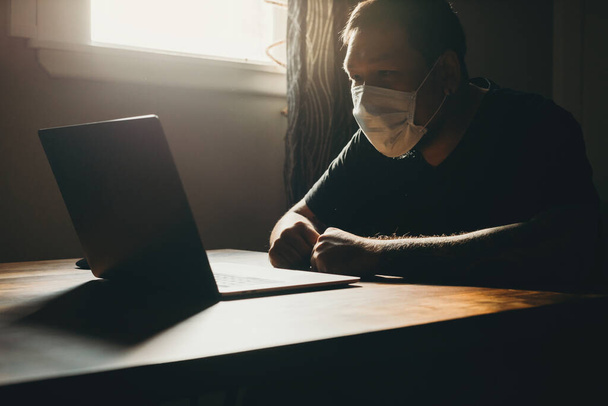 Un hombre asiático con máscara médica trabajando en el portátil en el área oscura. Gestos de tristeza
. - Foto, imagen