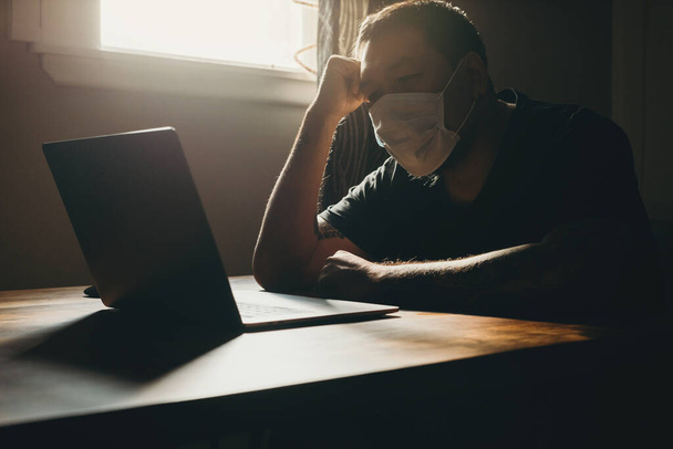 Азиат в медицинской маске работает на ноутбуке в тёмной зоне. Жест грусти
. - Фото, изображение