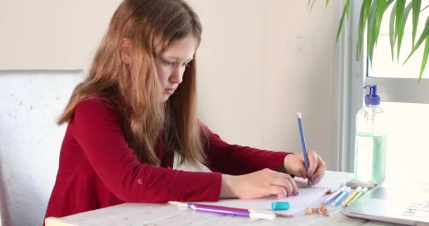 Distance learning online education. Schoolgirl studying at home with a laptop and doing school homework. Training books and colored felt-tip pens on the table, gel with alcohol 70 percent. Coronavirus - Imágenes, Vídeo