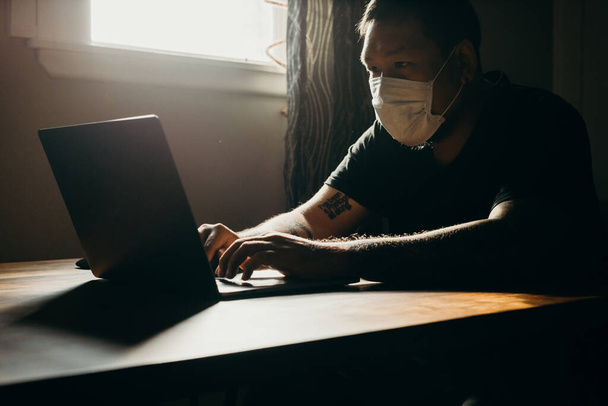 un asiático hombre en médico máscara trabajo en portátil en oscuro área
. - Foto, Imagen