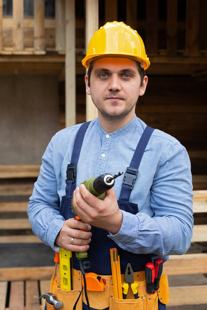 Young handsome carpenter with yellow hard hat and work equipment is holding an electric drill on wooden background - Foto, afbeelding