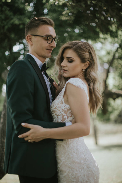 Portrait of a lovely bride and groom embrace, kiss and hug each other. Couple in love in the bosom of nature . Handsome groom and beautiful blonde bride posing in the forest. Modern dress and suit - Zdjęcie, obraz