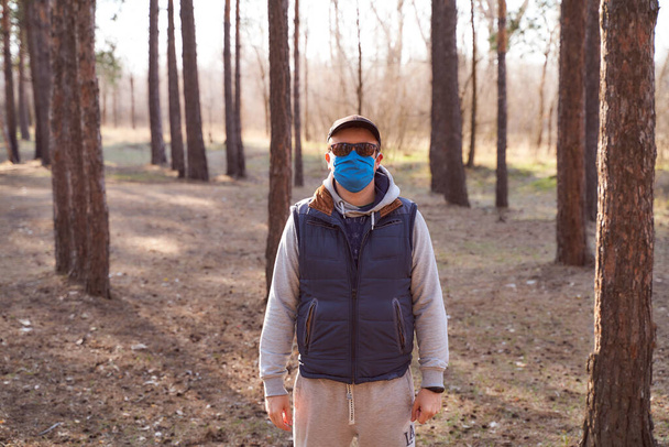 A lonely man in a tracksuit stands near the pines in a blue mask, cap and glasses - Fotografie, Obrázek