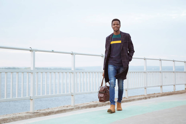 Black man in a coat, a pedagogue on the waterfront, against the background of the river, water, sea. Fashionable stylish look, image. Modern fashion. Holds a bag, a tourist in his hands. - Foto, Imagen