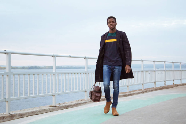 Black man in a coat, a pedagogue on the waterfront, against the background of the river, water, sea. Fashionable stylish look, image. Modern fashion. Holds a bag, a tourist in his hands. - Foto, afbeelding