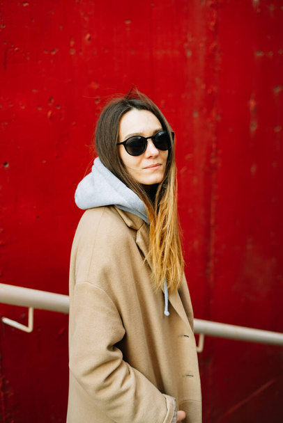 a young girl in a brown coat and glasses on the street, standing near a red wall. street style - Zdjęcie, obraz