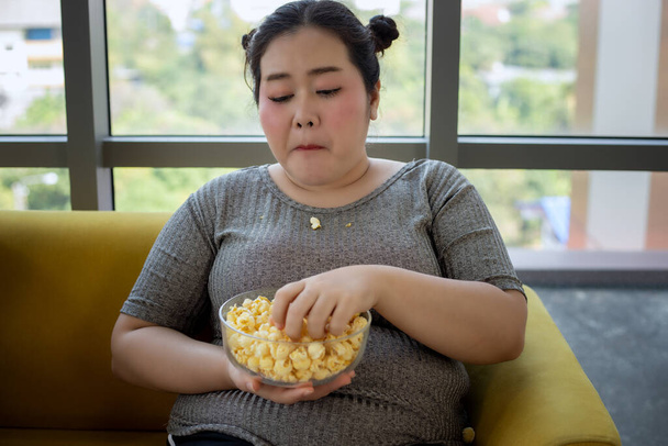 Ylipainoinen nainen ja aasialainen tyttö nauttivat ruoasta ja popcornista sohvalla kotona
 - Valokuva, kuva
