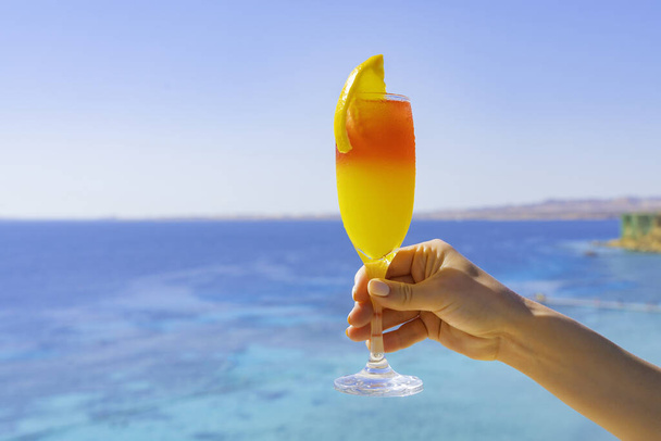 Meisje houdt in een hand verfrissende sap cocktail met sinaasappel. Blauwe zee op de achtergrond. Vakantie in de tropen. Zonnige gezonde ontspanning concept - Foto, afbeelding