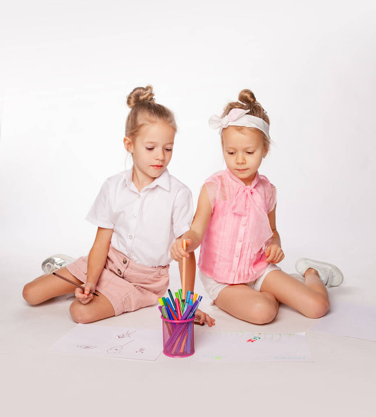two girls are sitting cross-legged and draws on paper with felt-tip pens.  - Zdjęcie, obraz