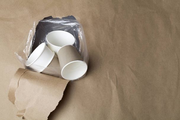 eco friendly disposable, compostable, recyclable paper cups in plastic bag through hole in brown paper background. land pollution concept. suffocates nature. copy space - Photo, Image