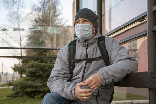 バスの停留所で医療マスクに座っている中年の男は、平均的な計画。概念:コロナウイルスからの市民の保護、市内の流行、人々のための安全を確保する手段. - 写真・画像
