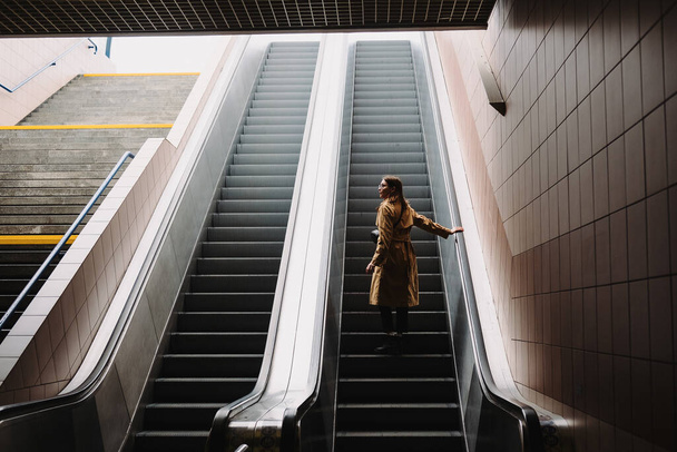 Młoda kobieta w płaszczu podnosi się na ruchomych schodach z metrem do miasta. - Zdjęcie, obraz