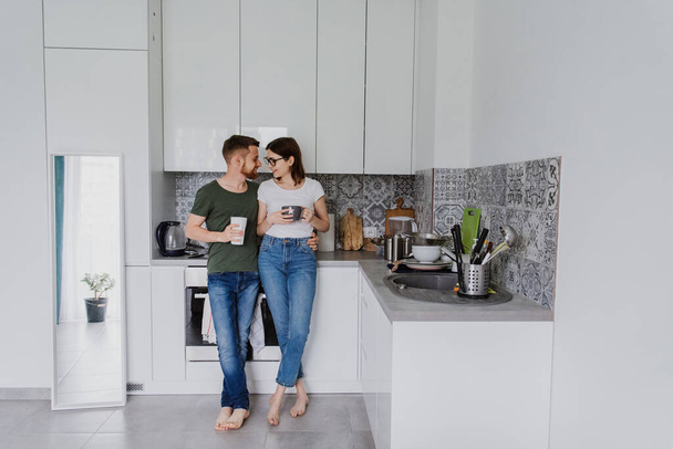 Νεαρός άντρας και γυναίκα γελάνε, αγκαλιάζουν και πίνουν τσάι στην κουζίνα. - Φωτογραφία, εικόνα