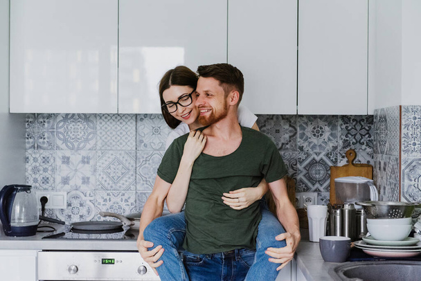 Νεαρός άντρας και γυναίκα γελούν και αγκαλιάζονται στην κουζίνα. Όμορφη γυναίκα αγκαλιάζει έναν άντρα από πίσω στην κουζίνα.. - Φωτογραφία, εικόνα