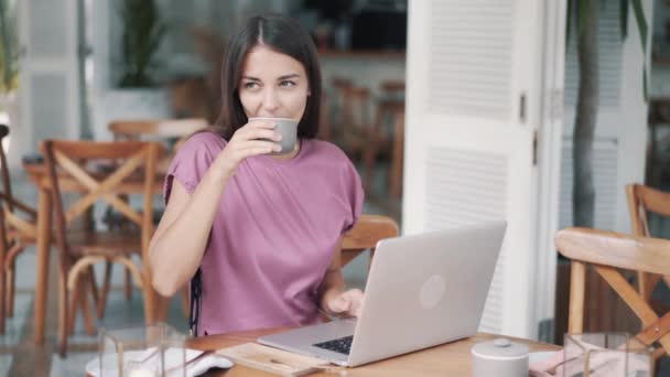 Beautiful young woman freelancer drinks coffee and uses laptop for work in cafe - Felvétel, videó