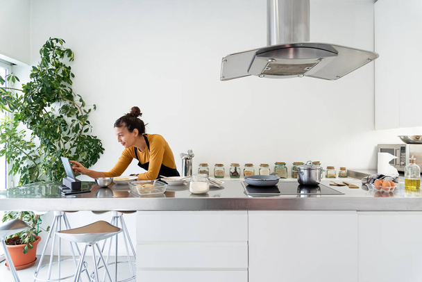 jonge brunette vrouw op zoek naar haar digitale tablet voor een recept op het web en koken in een moderne keuken - Foto, afbeelding