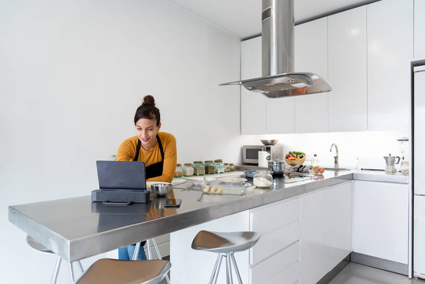 Young brunette woman looking at her digital tablet for a recipe on the web and cooking in a modern kitchen - Foto, Bild