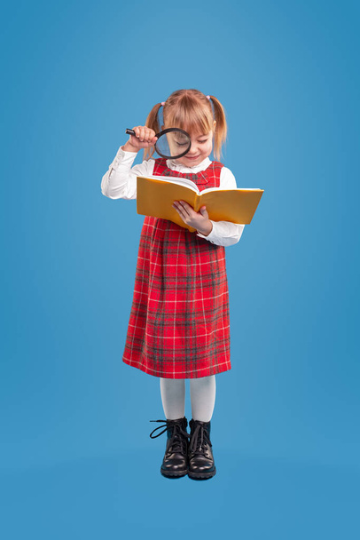 Cute little girl in red checkered school uniform reading textbook with magnifier while standing against blue background - Foto, Imagem