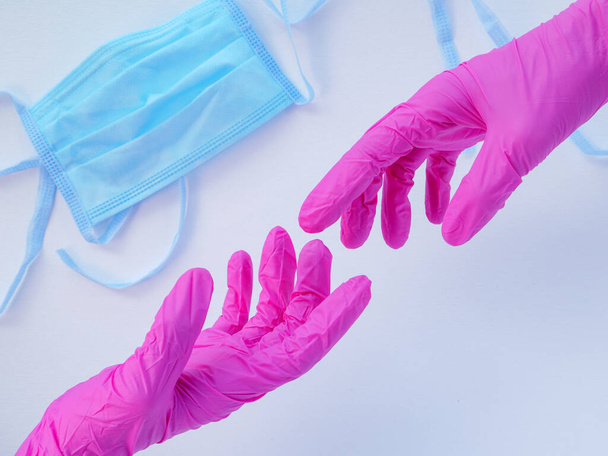 Twee handen in roze latex handschoenen die naar elkaar reiken. Blauw beschermend medisch gezichtsmasker op een witte achtergrond. Antivirale bescherming tijdens coronaviruspandemie. Ruimte voor tekst. - Foto, afbeelding