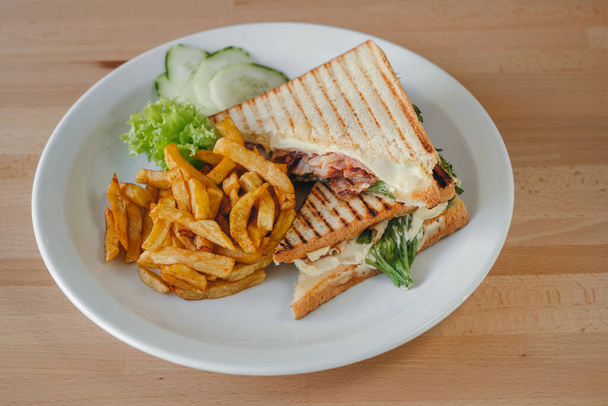 Szalonnás szendvics sült krumplival és zöldséggel, fehér tálcán, fa alapon. Étkezési idő. - Fotó, kép