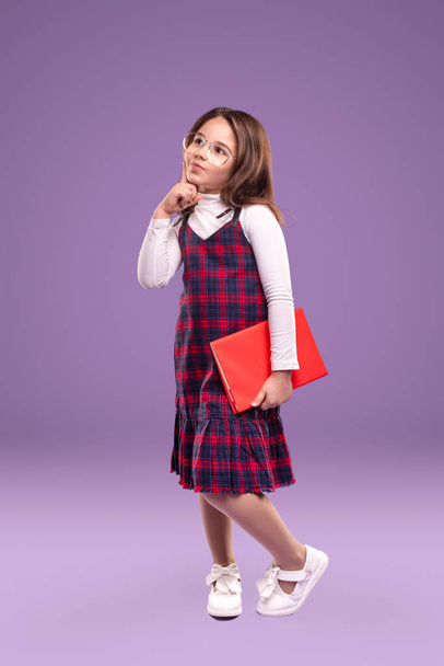 Cute clever pupil girl dressed in school uniform and eyewear holding red book and looking away thoughtfully while standing against purple background - Photo, Image