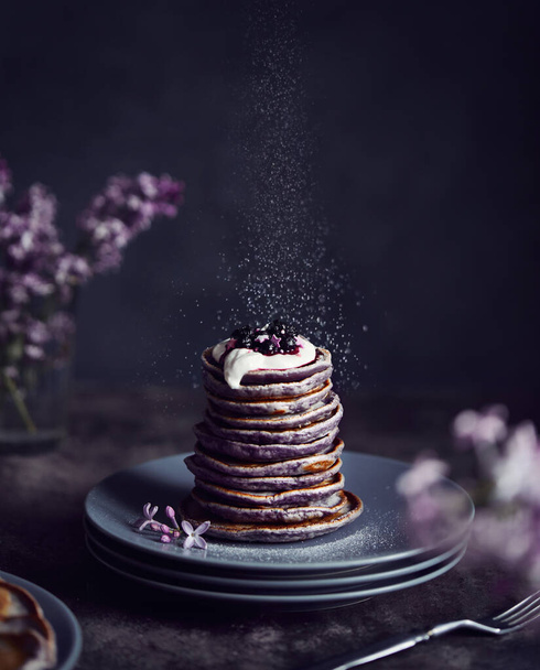 Violet pancakes with blueberries and cream around lilac flowers at dark atmospheric background in spring time - Foto, Imagem