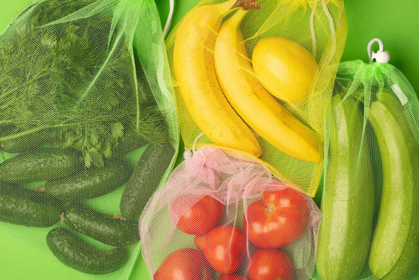 Крупный план фотографии огурцов, помидоров, бананов и цуккини в многоразовом прозрачном мешке, вид сверху на зеленый фон. Экологичные покупки с нулевыми отходами
. - Фото, изображение