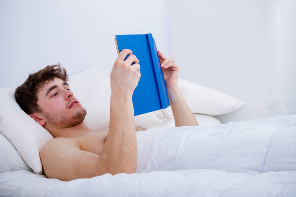 Egy fiatal felnőtt férfi, csupasz mellkassal az ágyban, könyvet olvasva. - Fotó, kép
