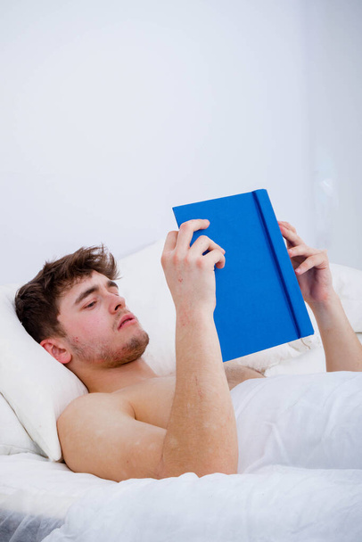 A young adult man, bare chested in bed reading a book - Foto, Imagen