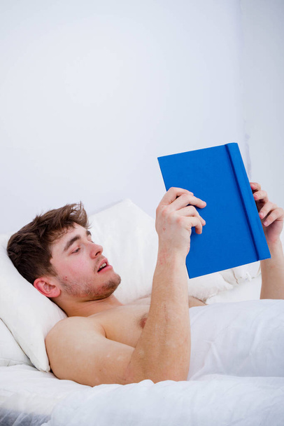 A young adult man, bare chested in bed reading a book - Фото, изображение