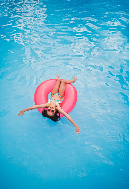 From above of cheerful little girl in swimwear and sunglasses floating on pink inflatable ring in swimming pool with blue water during summer holidays - Fotografie, Obrázek