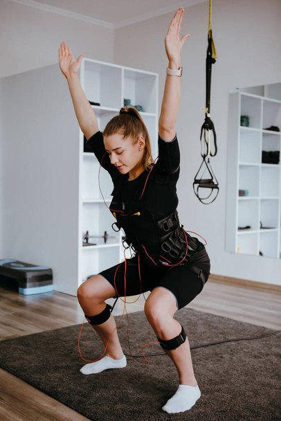 EMS training. Girl doing squats with her arms raised in suit with cables. - Fotó, kép