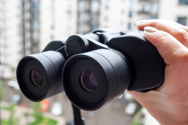 Hand holding black binoculars against a blurred background of a window with a view of the neighboring house for observing neighbors, environment or nature. Selective focus. Closeup view - Photo, Image