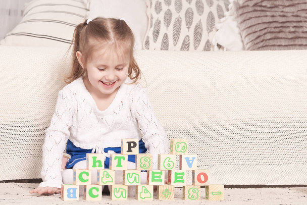a little three-year-old girl plays cubes on which letters and numbers are drawn, on the floor near the bed - Foto, afbeelding