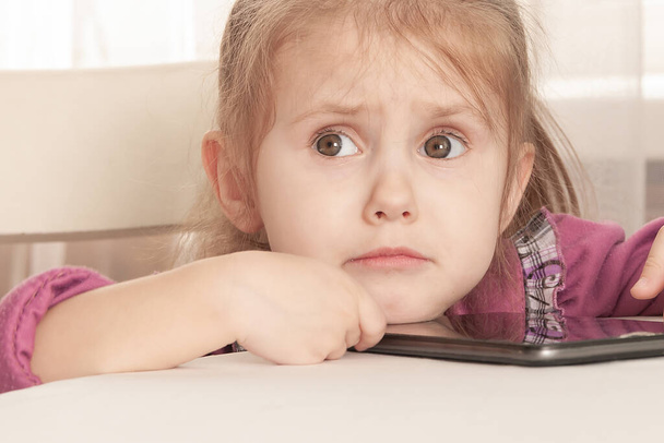displeased upset three year old girl in a purple dress sits at a table and plays with a tablet, resentment - Foto, afbeelding