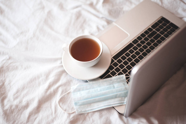 Kopje thee met medisch masker op open laptop in bed close-up. Thuis werken. Sociale afstand. Goede morgen. Quarantainetijd.  - Foto, afbeelding