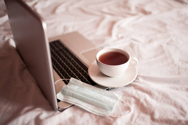 Itthon maradok. Egy csésze friss tea orvosi maszkkal nyitott laptopon. Karantén idő. Társadalmi távolság.  - Fotó, kép
