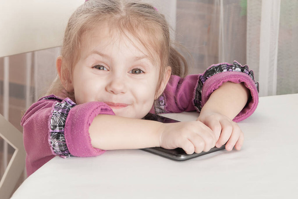 happy smiling three year old girl in a purple dress sits at a table and plays with a tablet, joy. - Foto, Imagen