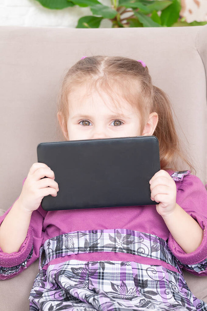 little girl in a purple dress sits in an armchair and plays with a tablet, a smile on her face - Foto, afbeelding