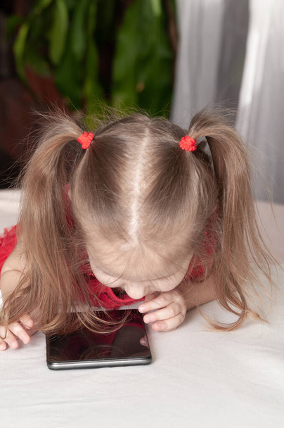 Kırmızı elbiseli küçük bir kız masada oturur ve tabletle oynar, düşünceli bir bakışla. - Fotoğraf, Görsel