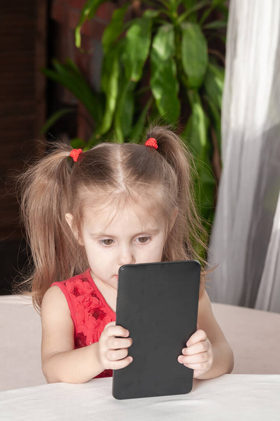 little girl in a red dress sits at a table and plays with a tablet, with a pensive look - Foto, afbeelding