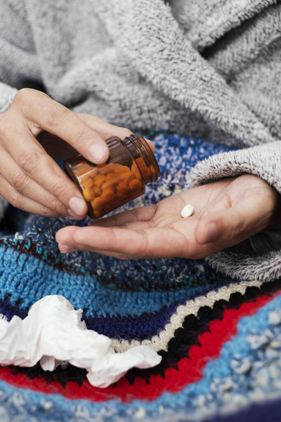 closeup of an ill caucasian man at home, wearing a fluffy gray house robe and wrapped in a colorful knitted blanket, about to take a pill from a bottle - Photo, Image