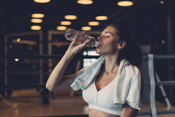 Πανέμορφη νεαρή γυναίκα με μια πετσέτα στους ώμους της πίνοντας νερό από ένα μπουκάλι στο γυμναστήριο - Φωτογραφία, εικόνα