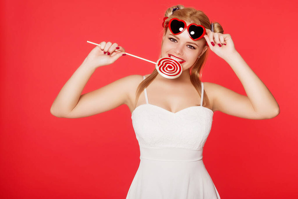 Beautiful blond woman dressed in an anime style is holding a lollipop. Glasses of the heart. Snow white broad smile. Copy space. - Φωτογραφία, εικόνα