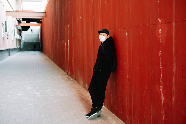 a young man in black sportswear and a baseball cap with a medical mask on his face near the red wall. protection from the virus - Foto, afbeelding